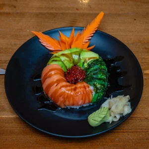 Sushi donut with salmon, avocado, seaweed salad, and fish roe on rice, served with wasabi, pickled ginger, and soy sauce drizzle on a black plate.